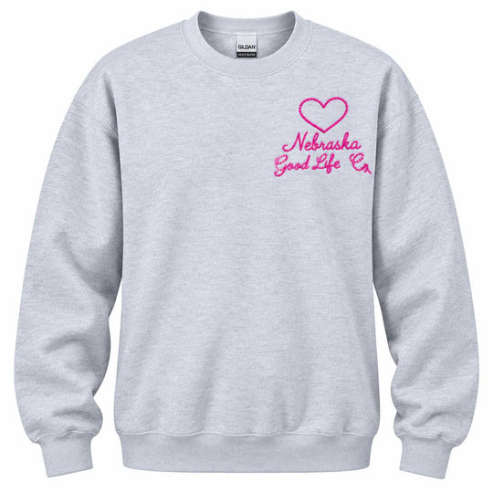 A light heather gray crewneck sweatshirt featuring a pink embroidered heart and the text 'Nebraska Good Life Co' on the left chest, displayed on a clean white background.