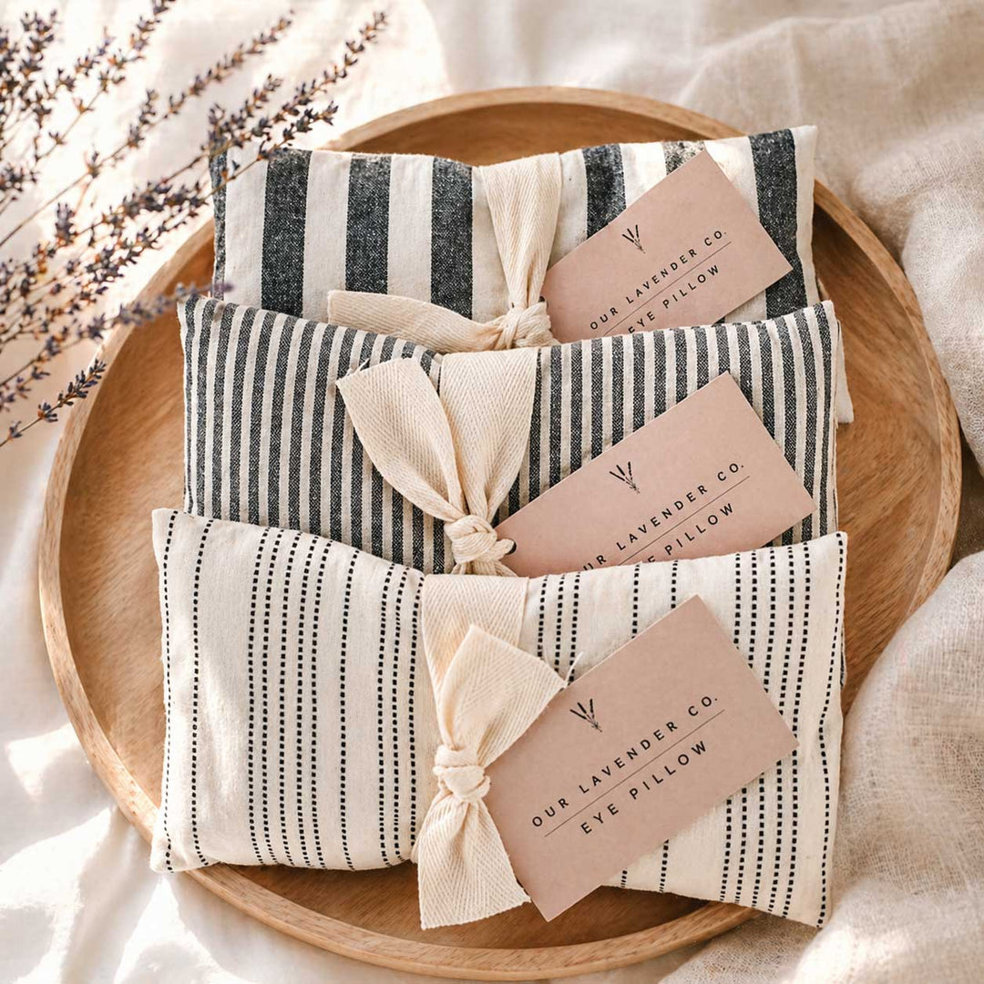 Striped lavender eye pillows with tags on a wooden plate, surrounded by lavender flowers.