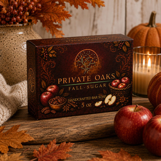 A boxed bar of soap labeled Private Oaks Fall Sugar sits on a rustic wooden surface surrounded by fall-themed props including apples, leaves, pumpkins, and a lit candle.