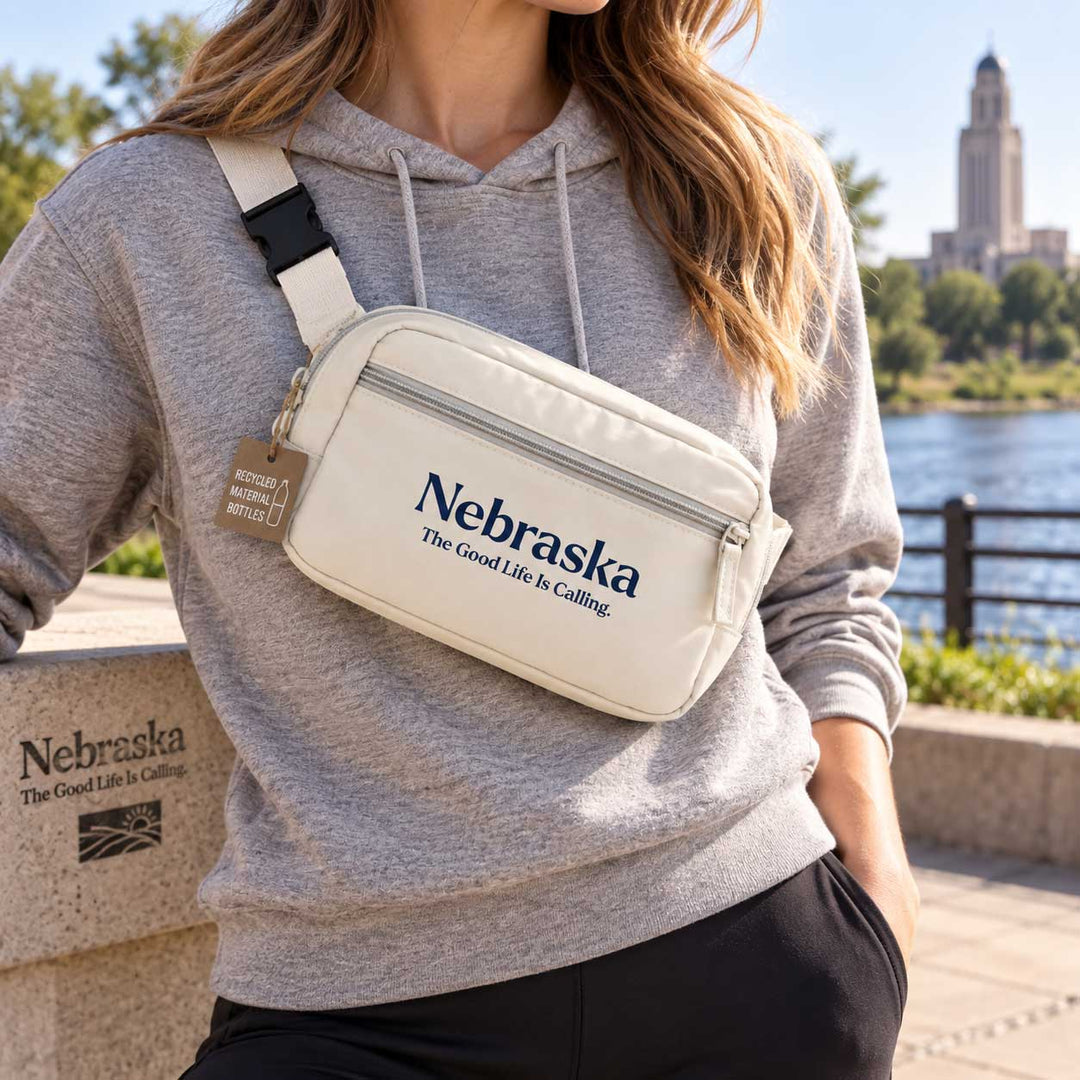 A woman wearing a gray hoodie and black pants carries a cream-colored crossbody bag with the text 'Nebraska The Good Life Is Calling' printed on the front. The bag has a white strap with a black buckle and a small brown hanging tag.