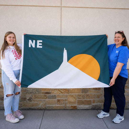 Two women holding a dark green flag with a white mountain, a yellow sun, and the letters 'NE' in white, standing outdoors against a beige wall and stone base.