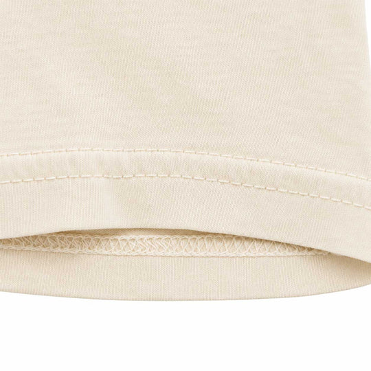 Close-up detail of a cream-colored t-shirt hem, showing the ribbed knit fabric, double stitching, and folded edge against a white background.