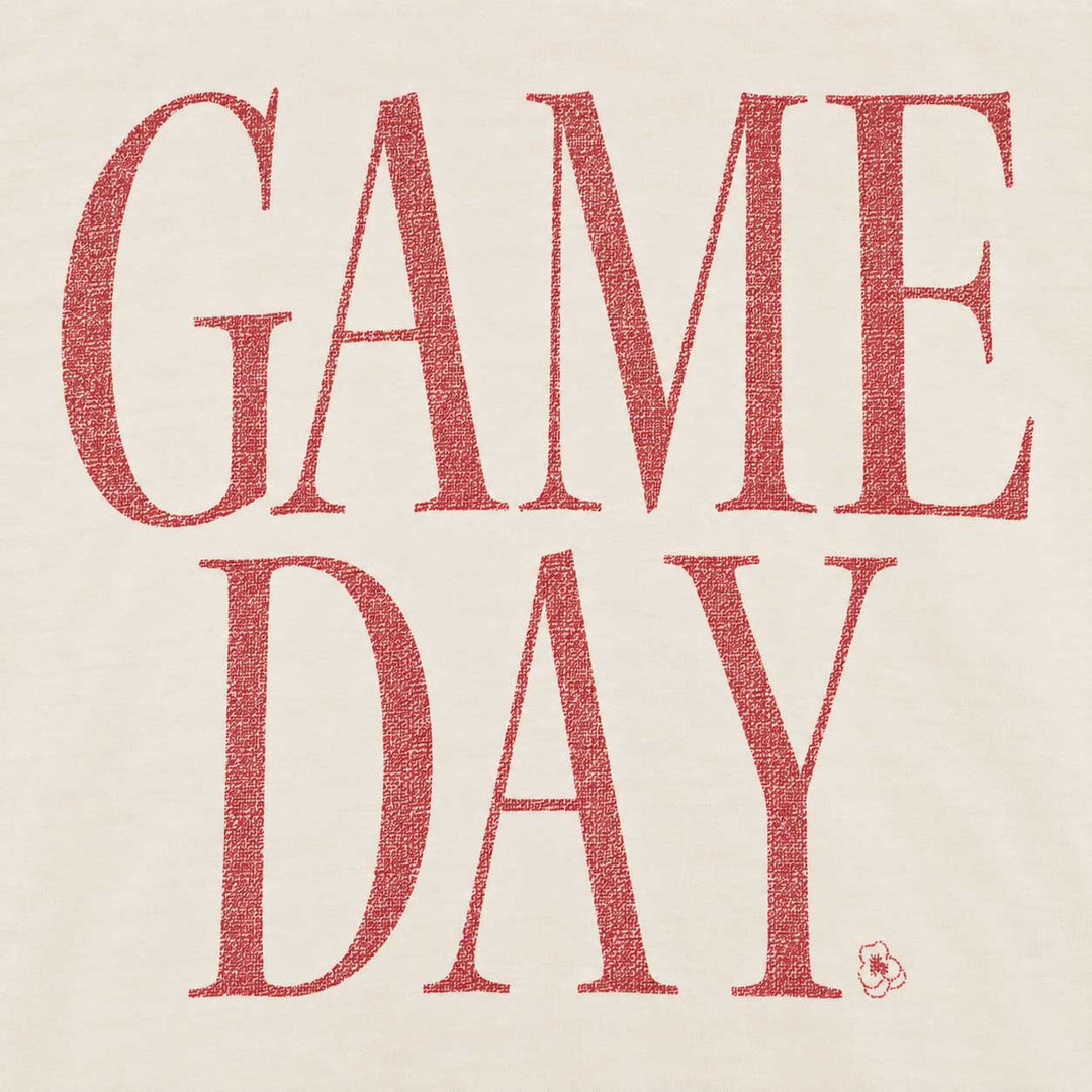 A close-up of a light beige fabric surface with large red text reading “GAME DAY.” in a textured serif typeface, plus a small red logo mark near the lower right of the lettering.
