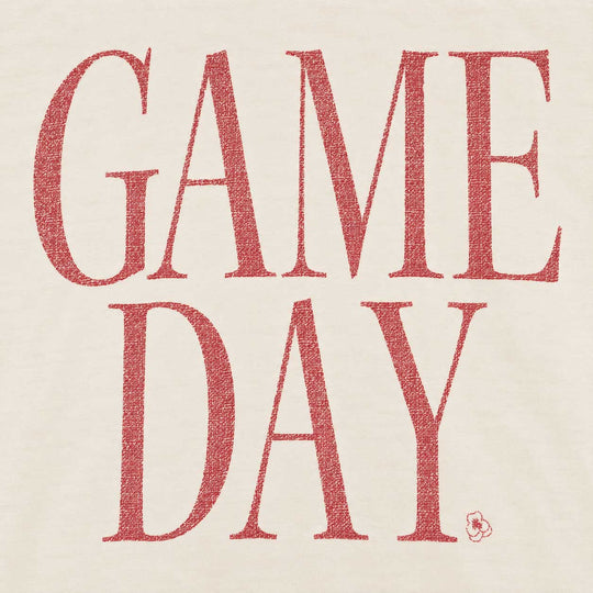 A close-up of a light beige fabric surface with large red text reading “GAME DAY.” in a textured serif typeface, plus a small red logo mark near the lower right of the lettering.