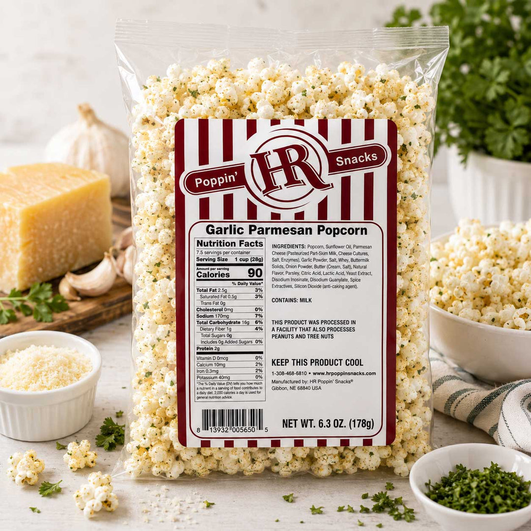 A bag of Poppin' R Snacks Garlic Parmesan Popcorn stands on a light stone surface with bowls of cheese, parsley, and extra popcorn around it. The package is clear plastic with a large front label showing the brand and product name, and the popcorn inside is pale and creamy.