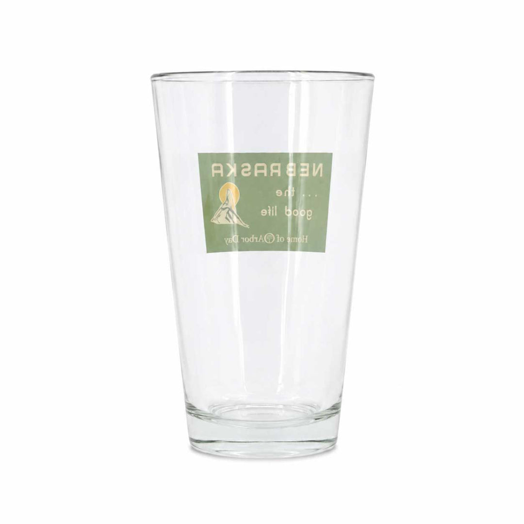 A clear glass pint glass with a green rectangular label on the front featuring a small mountain illustration and mirrored text, shown on a plain white background.