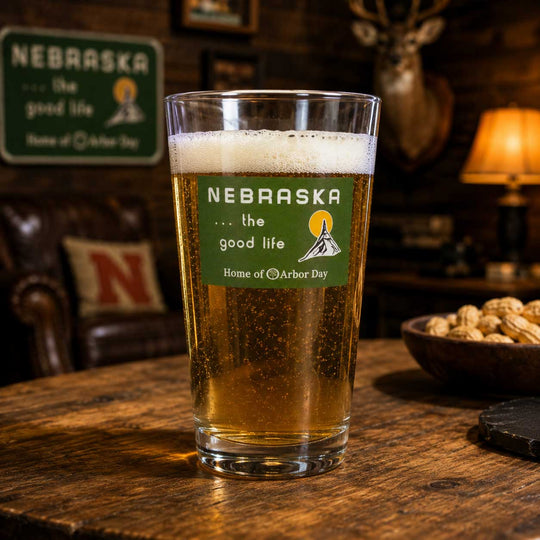 A clear pint glass filled with golden beer and foam, printed with a green Nebraska state graphic and text, set on a wooden table in a warmly lit bar or lounge with a lamp, peanuts, and leather seating in the background.