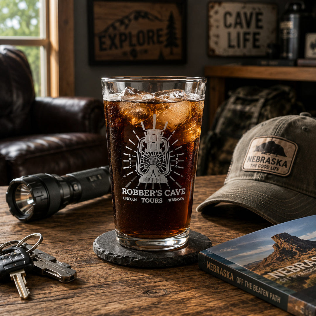 A clear glass drinking cup filled with iced dark cola sits on a coaster on a wooden table in a rustic room. The cup is etched with a mountain and the words 'Robber's Cave' and 'Lincoln Tours Nebraska.