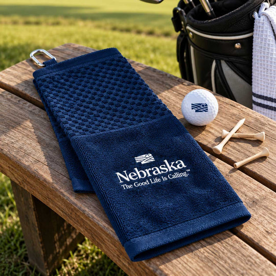 A navy blue textured golf towel with white Nebraska branding is laid on a wooden bench outdoors, with a golf bag, golf ball, and tees nearby.