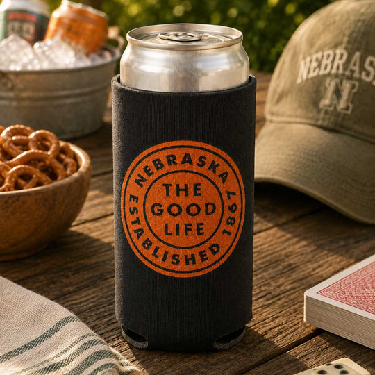 A black neoprene can cooler sleeve with an orange circular 'Nebraska The Good Life Established 1961' print, holding a silver aluminum can on a rustic wooden table surrounded by beer, a hat, and pretzels.