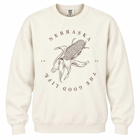 An off-white crewneck sweatshirt featuring a dark brown graphic of a corn cob with a person dancing in front of it, surrounded by the text 'NEBRASKA THE GOOD LIFE 1992'.