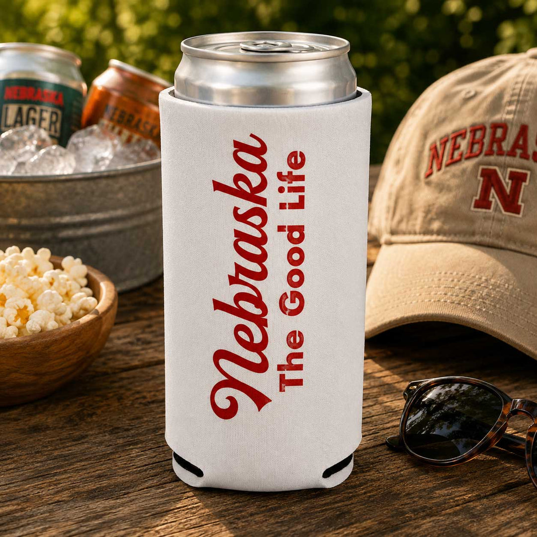 A white can cooler sleeve with red 'Nebraska The Good Life' text, holding a silver can on a wooden table with a cap, sunglasses, popcorn, and ice bucket in a sunny outdoor setting.