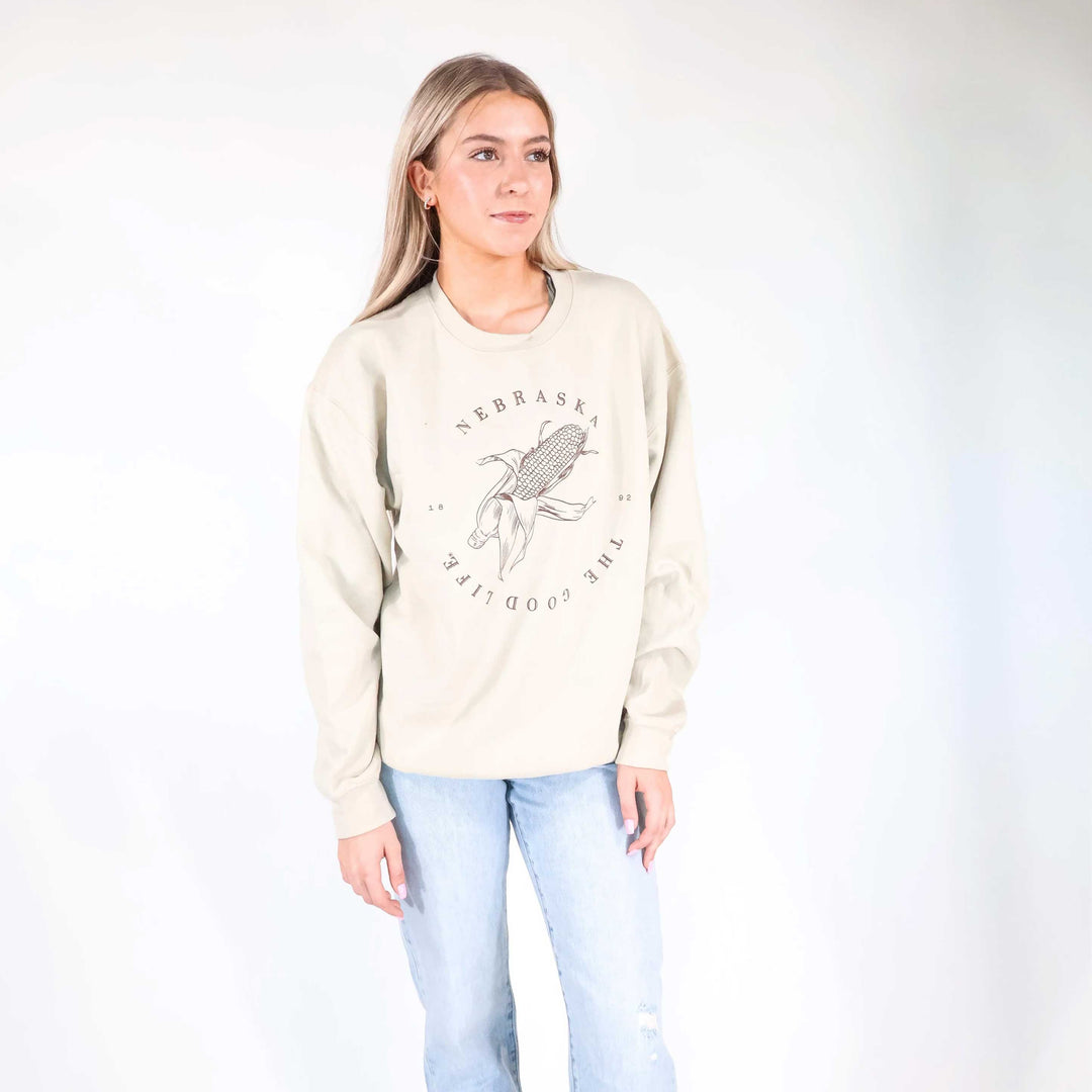 A woman modeling a cream-colored crewneck sweatshirt with a black graphic of a corn cob and the text 'NEBRASKA' and 'THE GOOD LIFE' on the front, paired with light blue jeans against a white studio background.