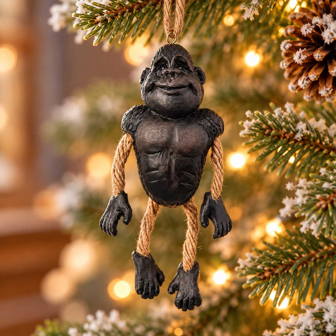 A novelty gorilla Christmas ornament hanging from a tree branch. The ornament is sculpted like a muscular black gorilla with rope arms and legs, set against a blurred background of a decorated Christmas tree with warm lights.
