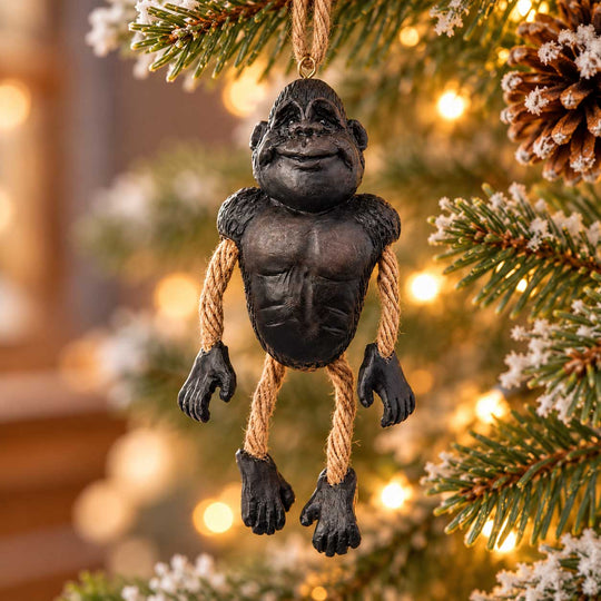 A novelty gorilla Christmas ornament hanging from a tree branch. The ornament is sculpted like a muscular black gorilla with rope arms and legs, set against a blurred background of a decorated Christmas tree with warm lights.