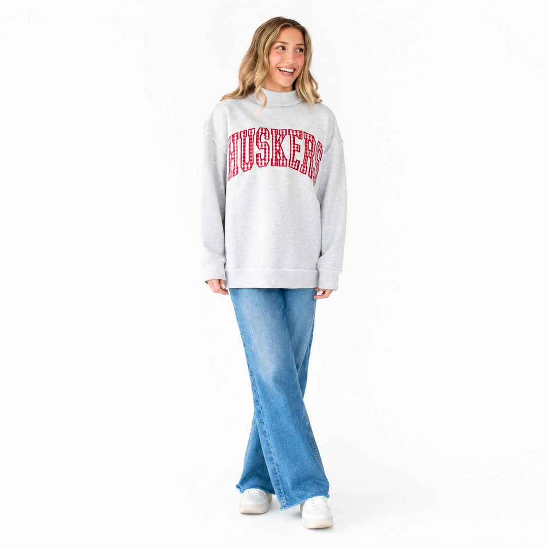 A woman modeling a light gray sweatshirt with 'HUSKERS' printed in red arched lettering across the chest, paired with light blue jeans and white sneakers, against a plain white background.