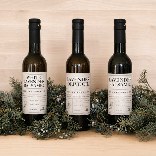 Three bottles of lavender-themed olive oil and balsamic vinegar on a wooden surface with greenery.