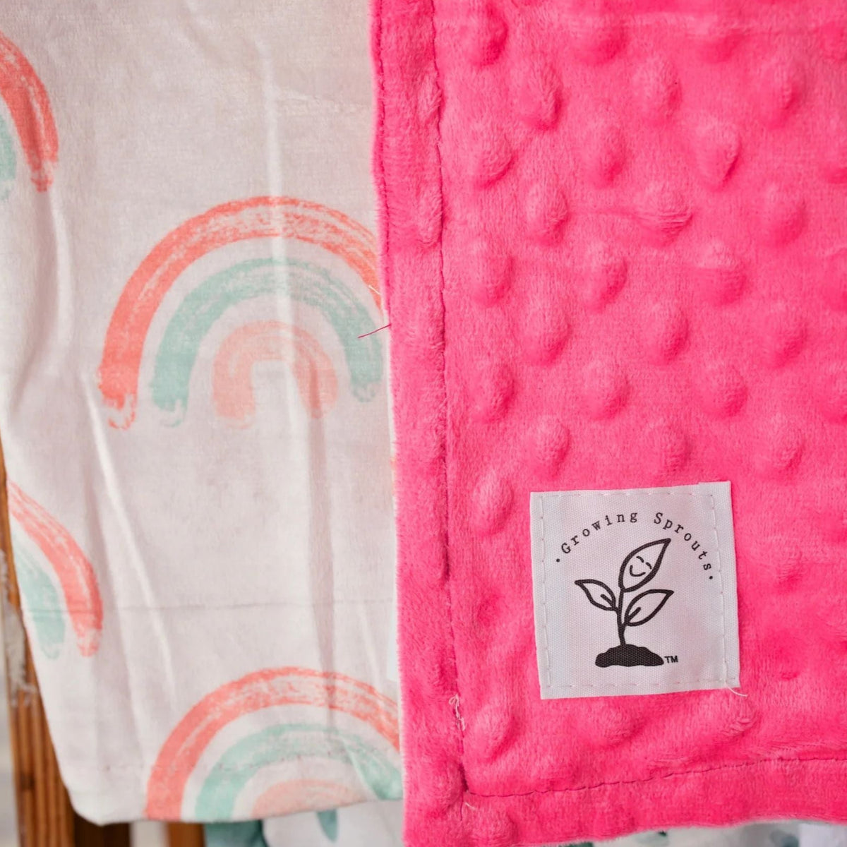 Pink textured blanket with a white label featuring a tree design, next to a white blanket with rainbow patterns.