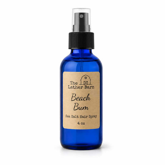 A 4 oz cobalt blue glass spray bottle of Beach Bum Sea Salt Hair Spray by The Lather Barn, featuring a kraft paper label and a black spray nozzle on a white background.