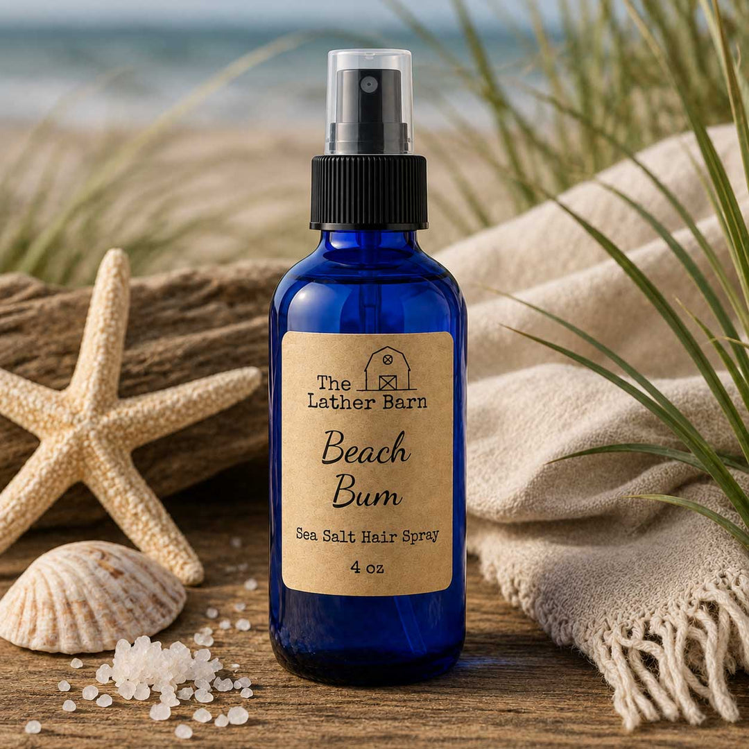 A 4 oz cobalt blue glass spray bottle of 'The Lather Barn' Beach Bum Sea Salt Hair Spray, styled on a wooden surface with a starfish, seashells, coarse sea salt, and a beige fringed towel against a blurred beach background.