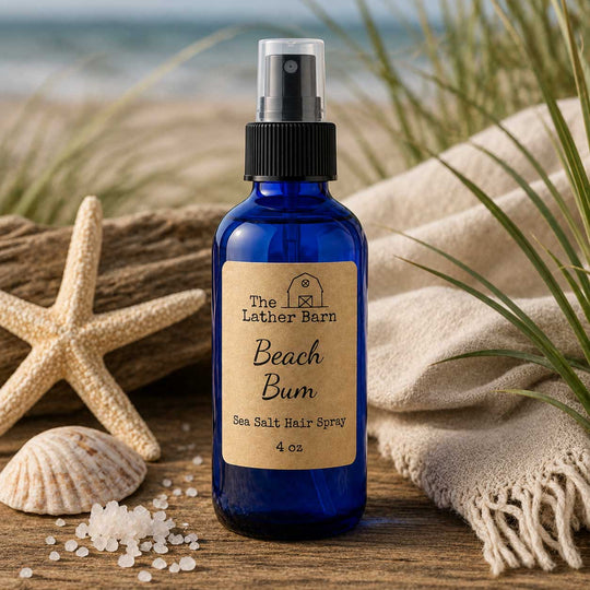 A 4 oz cobalt blue glass spray bottle of 'The Lather Barn' Beach Bum Sea Salt Hair Spray, styled on a wooden surface with a starfish, seashells, coarse sea salt, and a beige fringed towel against a blurred beach background.