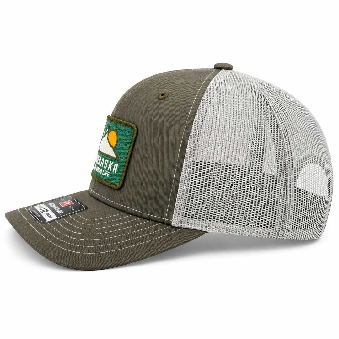An olive green and white trucker hat with a mesh back, featuring a green 'Alaska Native Life' patch on the front and a size tag on the brim.