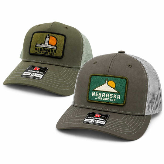 Two Nebraska trucker hats on a white background, one in olive green and one in gray. Both feature a rectangular patch with the Nebraska state silhouette and sun, and a visible Richardson 112 size 72 size label on the brim.