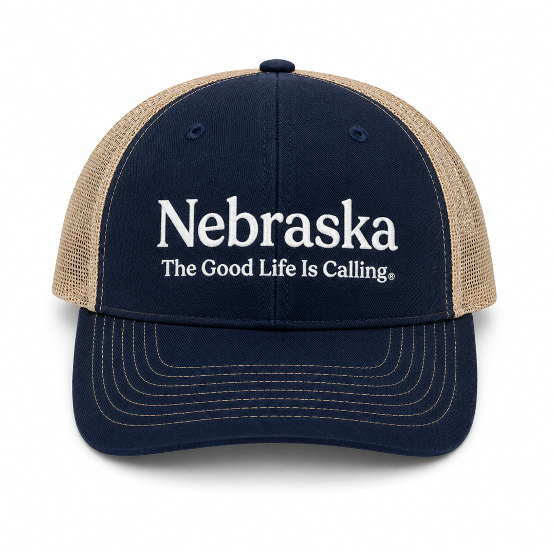 A navy blue and tan trucker hat with white embroidered text reading 'Nebraska The Good Life Is Calling' on the front, shown against a plain white background.