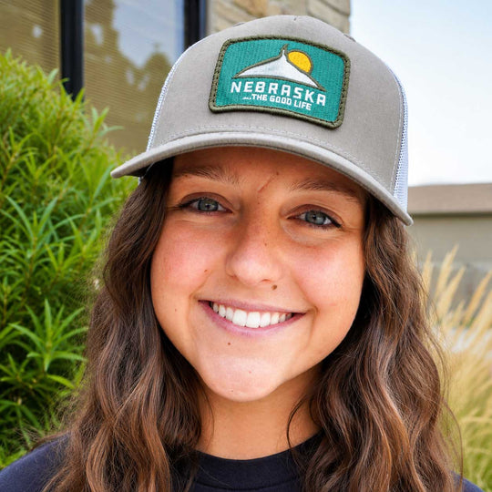 A smiling young woman wearing a gray trucker-style baseball cap with a green Nebraska patch on the front, photographed outdoors with plants and a building in the background.