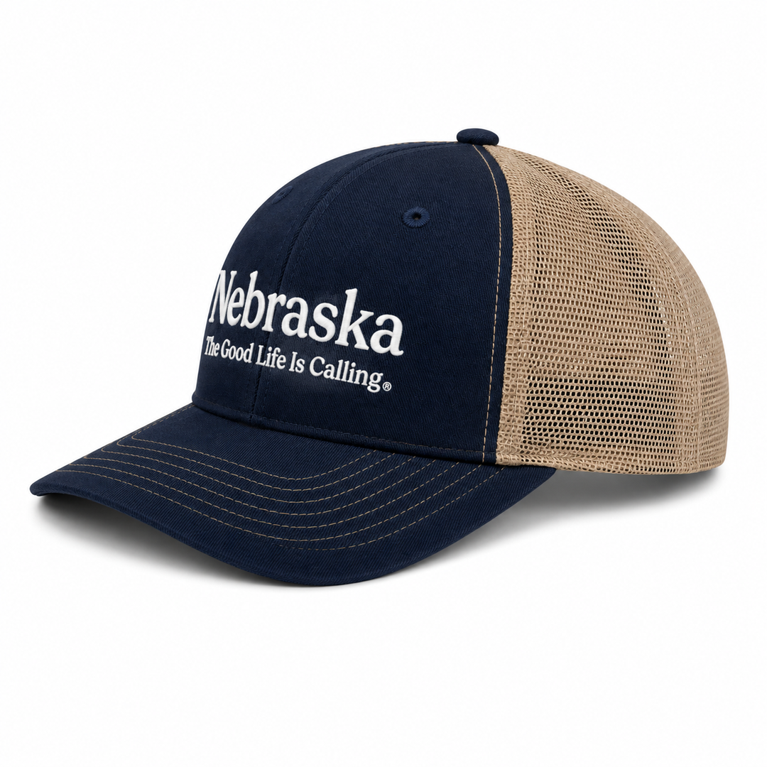 A navy blue and tan trucker hat with white embroidered text reading 'Nebraska The Good Life Is Calling' on the front panel, shown on a clean white background.