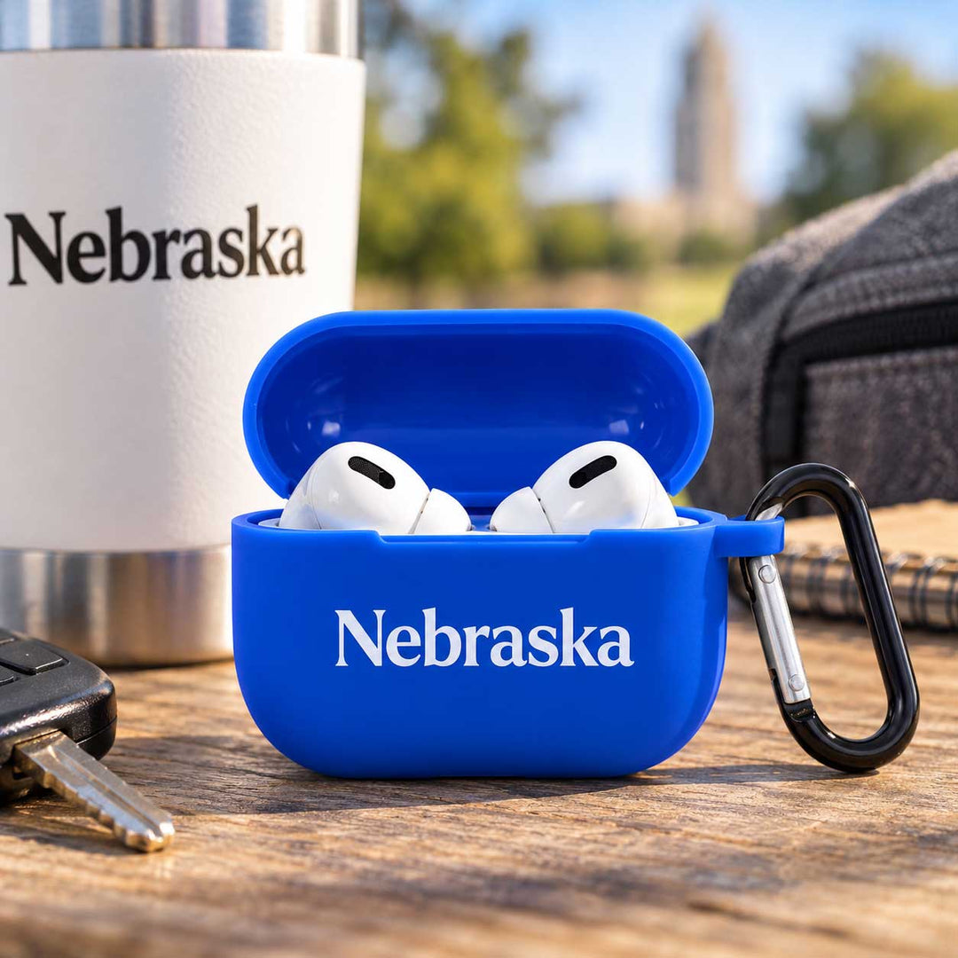 A bright blue AirPods case with 'Nebraska' printed in white, shown open with white AirPods inside on a wooden table. A white insulated tumbler with 'Nebraska' is blurred in the background, along with a car key and a gray backpack.