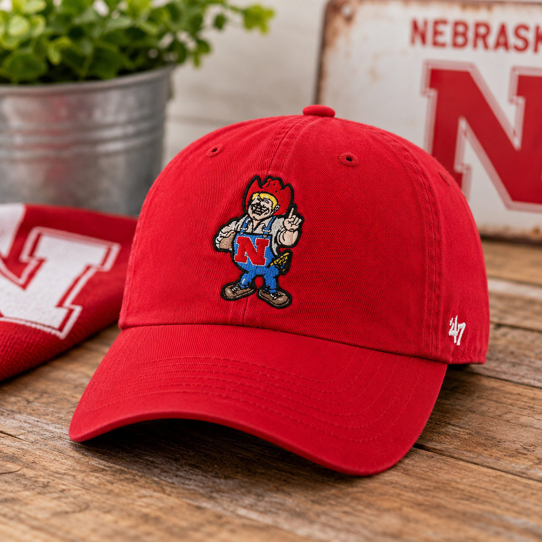 A red baseball cap with an embroidered Nebraska Cornhuskers mascot logo on the front, displayed on a rustic wooden surface with a matching red and white cap and a potted plant in the background.