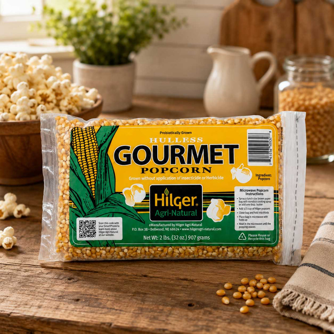 A bag of Hilger Gourmet Popcorn on a rustic wooden table with scattered popcorn kernels, loose corn kernels, and a small cream pitcher in the background.