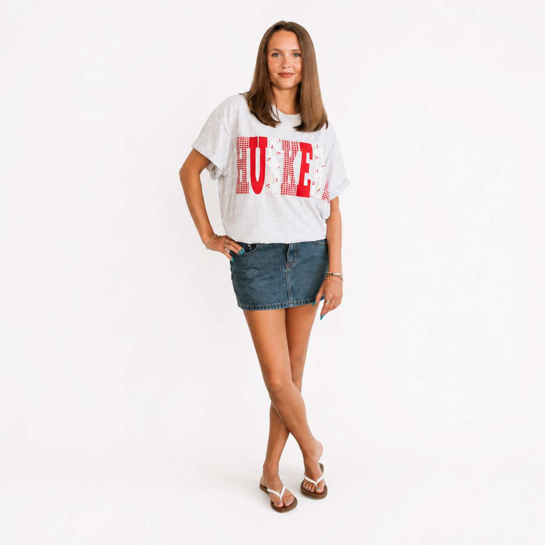 A woman modeling a white graphic t-shirt with a distressed red 'HUE' print, paired with a blue denim mini skirt and white slide sandals, against a plain white studio background.