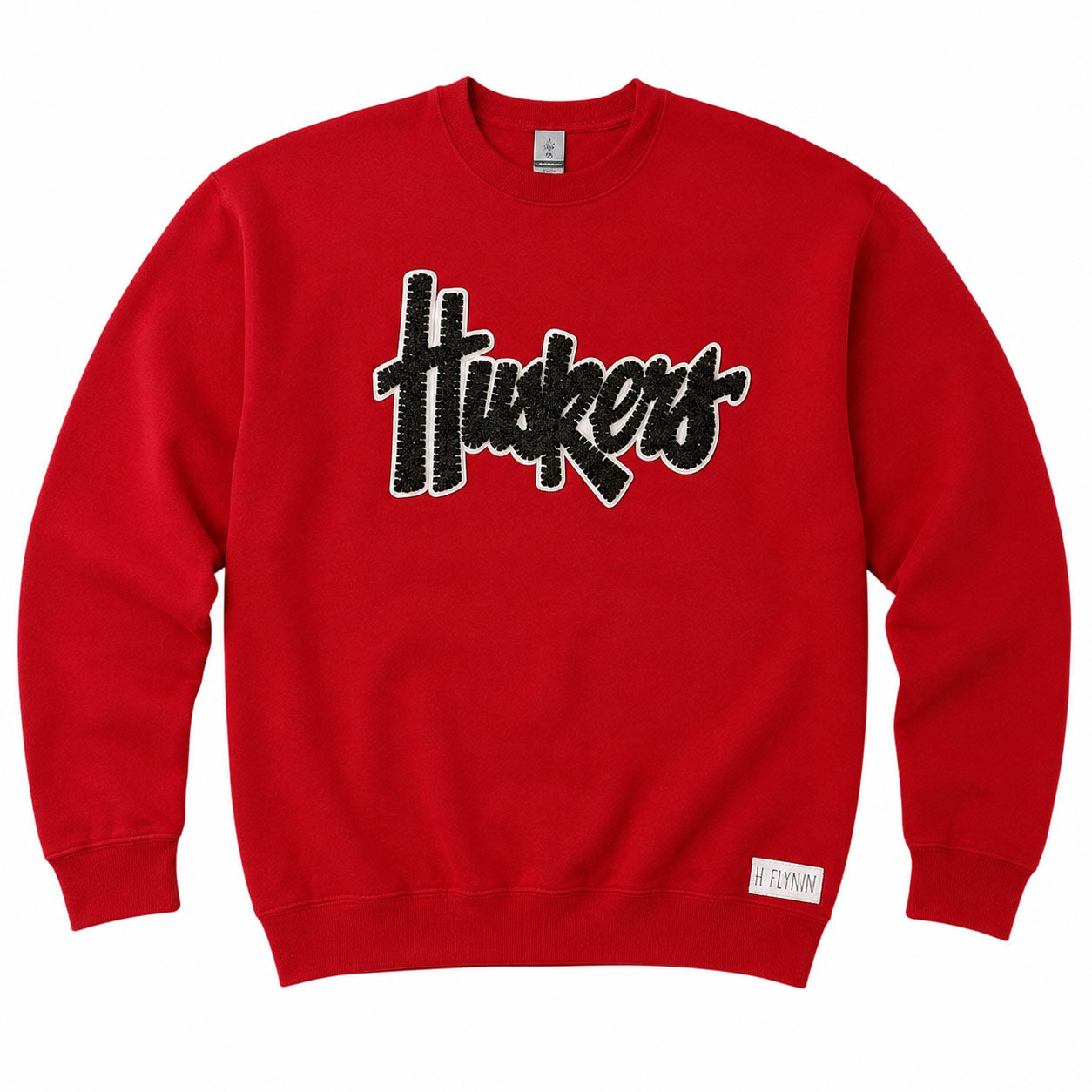A red crewneck sweatshirt featuring a black and white textured 'Huskers' graphic across the chest, with a small white tag near the bottom hem.