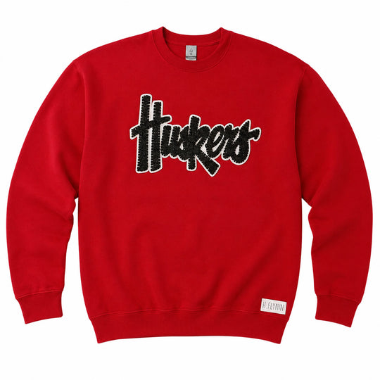 A red crewneck sweatshirt featuring a black and white textured 'Huskers' graphic across the chest, with a small white tag near the bottom hem.