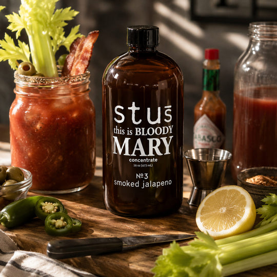 An amber glass bottle labeled 'stus this is BLOODY MARY concentrate' surrounded by Bloody Mary ingredients and garnishes on a wooden cutting board, including celery, jalapeños, a lemon slice, a metal jigger, and a jarred drink.