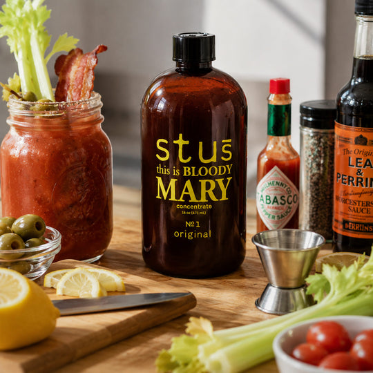A dark amber glass bottle with a black cap and bold yellow text reading 'st us this is BLOODY MARY concentrate' stands on a wooden surface. Surrounding it are food props including celery, tomatoes, olives, lemon slices, a cutting board, and other condiment bottles, suggesting a themed drink or sauce product in a kitchen-like setting.