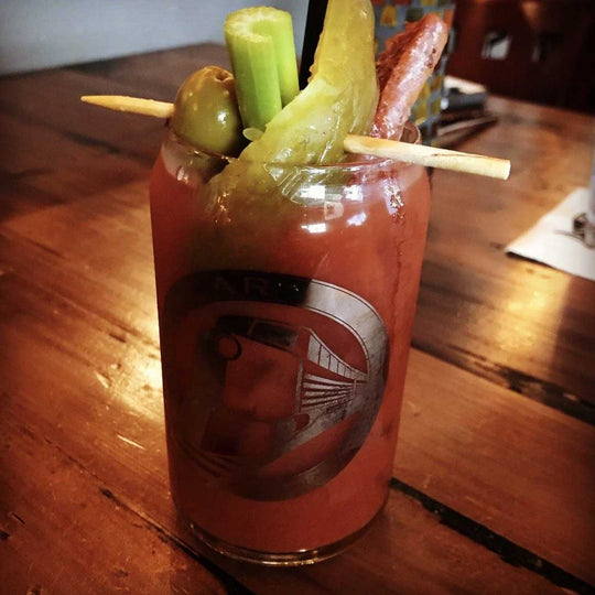 A Bloody Mary cocktail in a clear glass with a circular logo decal, garnished with celery, a pickle spear, an olive, and a hot dog stick on a wooden table.