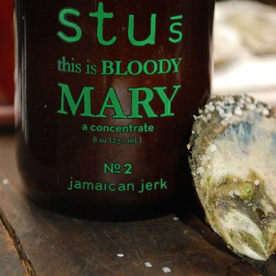 A close-up of a dark brown bottle labeled 'stus this is BLOODY MARY a concentrate 8 oz (237 mL)' with green text, placed on a wooden surface next to an oyster shell.