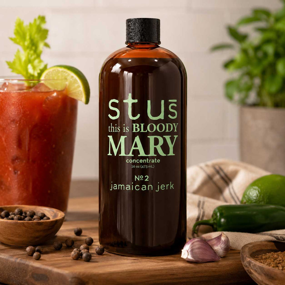 A bottle of Stus Bloody Mary concentrate (Jamaican jerk) stands on a wooden cutting board surrounded by ingredients like a Bloody Mary cocktail, garlic, jalapeño, and peppercorns. The background features a blurred kitchen setting with a potted plant.