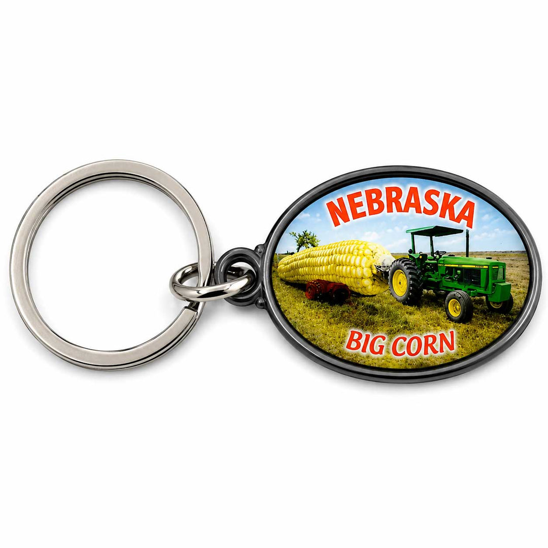 An oval keychain featuring a colorful printed design of a green tractor pulling a large corn cob in a field, with the words 'NEBRASKA' at the top and 'BIG CORN' at the bottom. The keychain has a silver metal split ring and key ring.