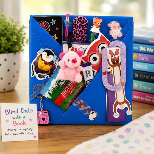 A blue blind date with a book mystery box organizer on a wooden table, filled with various colorful book-themed accessories, stickers, and a small pink plush toy. A card reading 'Blind Date with a Book' is placed in the foreground, with books and a plant blurred in the background.