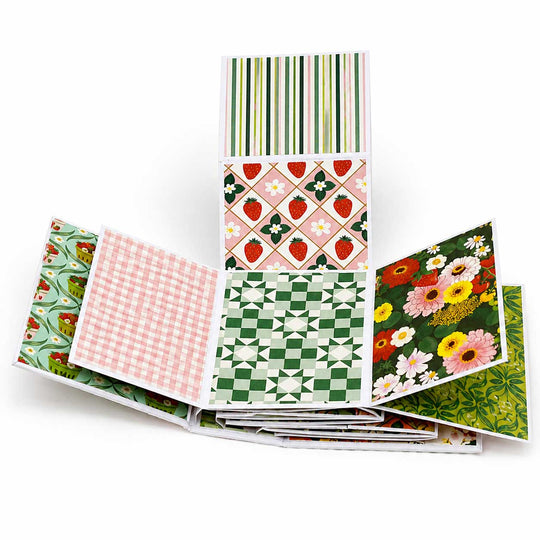 A stack of patterned paper sheets fanned out on a white background, showing various designs including stripes, strawberries, gingham, checkerboard, and floral patterns.