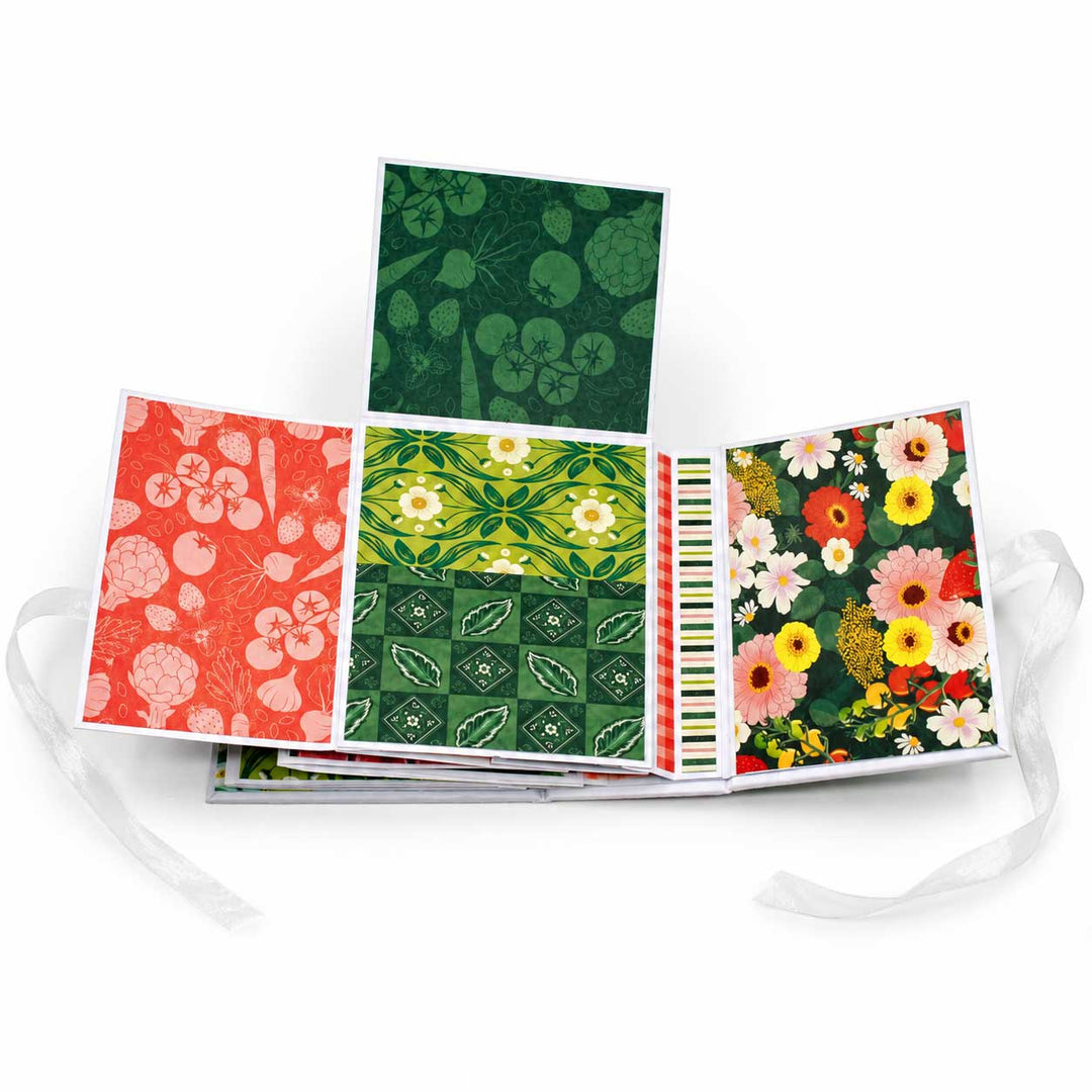 A stack of folded patterned cards or art prints fanned out on a white background, showing floral and botanical designs in red, green, yellow, and white, with white ribbon ties extending from the sides.