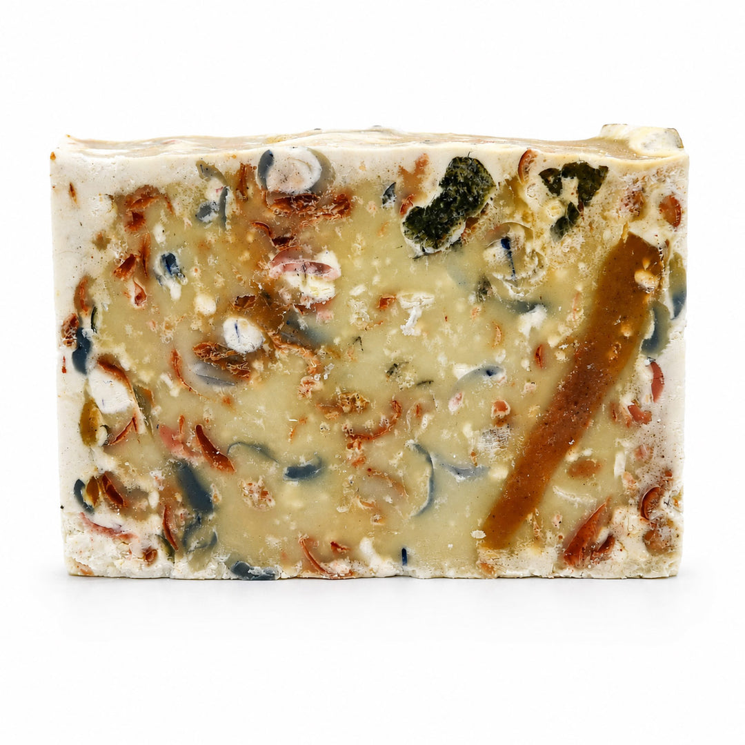 A rectangular bar of handmade soap with a textured, speckled cream and beige base, embedded with various natural ingredients including brown pieces, dark green bits, and small blue-gray inclusions, shown on a plain white background.