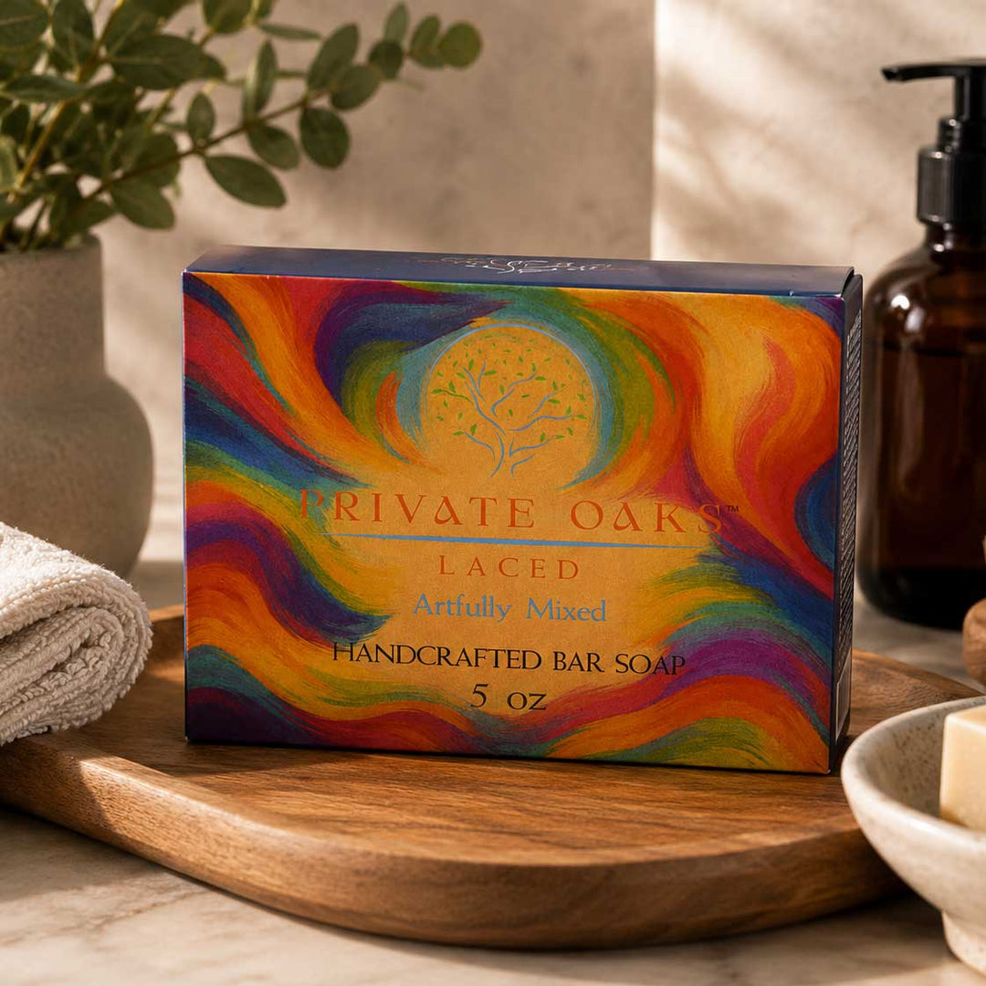 A colorful box of Private Oaks Laced Handcrafted Bar Soap sits on a wooden tray in a bathroom setting with a towel, soap dispenser, and marble surface.