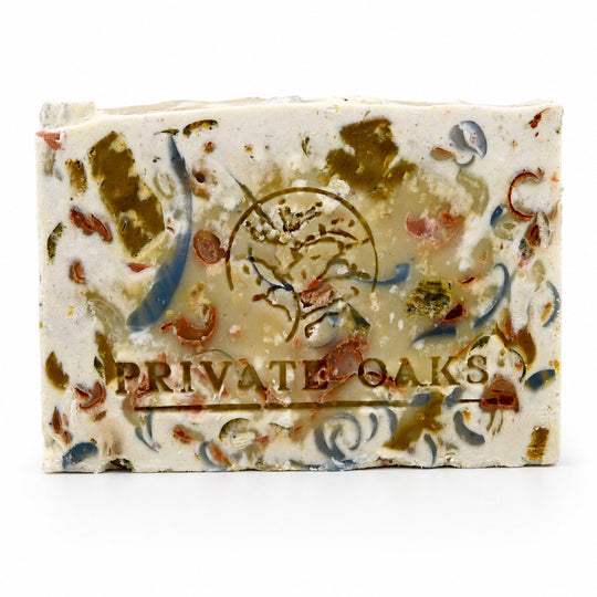 A rectangular bar of handmade soap with a cream-colored base, decorated with embedded brown, blue, and red botanical pieces. The bar features an embossed circular logo and the text 'PRIVATE OAKS' printed on the front, set against a plain white background.