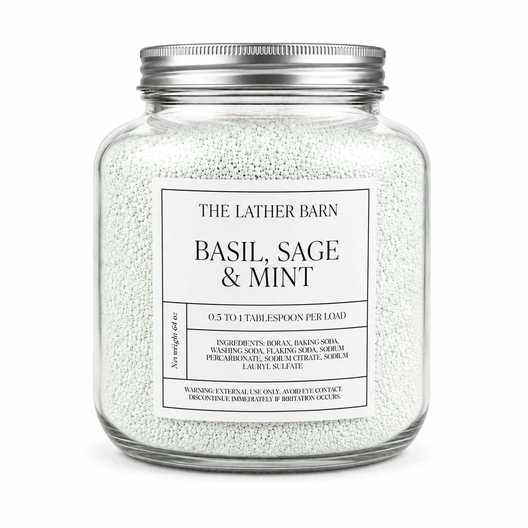 A clear glass jar filled with white bath salts or scrub beads, sealed with a silver metal lid, and labeled 'THE LATHER BARN BASIL, SAGE & MINT' on a white background.