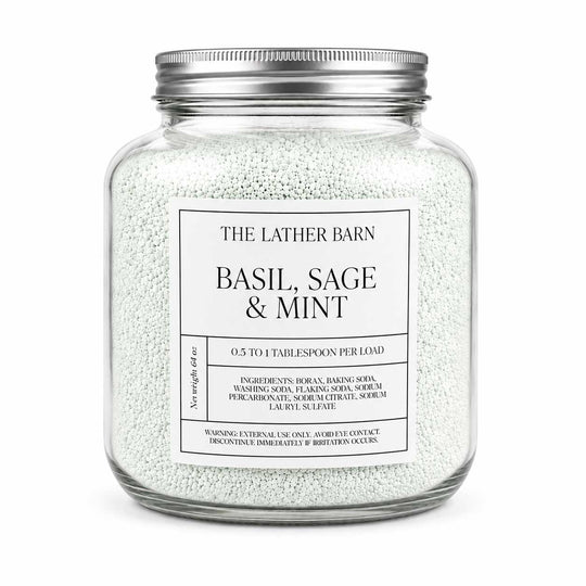 A clear glass jar filled with white bath salts or scrub beads, sealed with a silver metal lid, and labeled 'THE LATHER BARN BASIL, SAGE & MINT' on a white background.